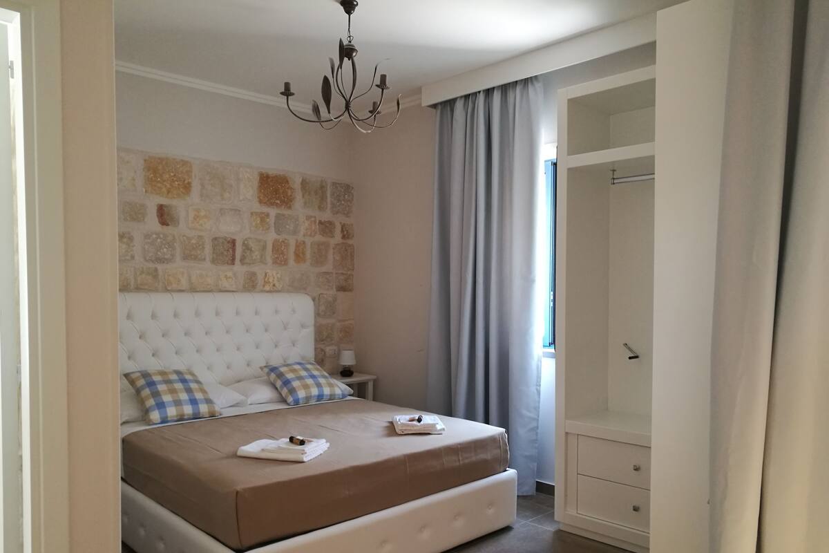 A bedroom with a large bed, a stone wall, a chandelier, a window with curtains, and a wardrobe.