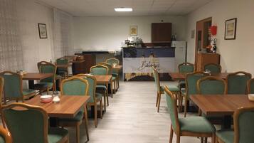 Un comedor con mesas de madera y sillas tapizadas en verde, una zona de cocina con refrigerador y armarios, y un letrero en la pared con el nombre "Bakerberger".
