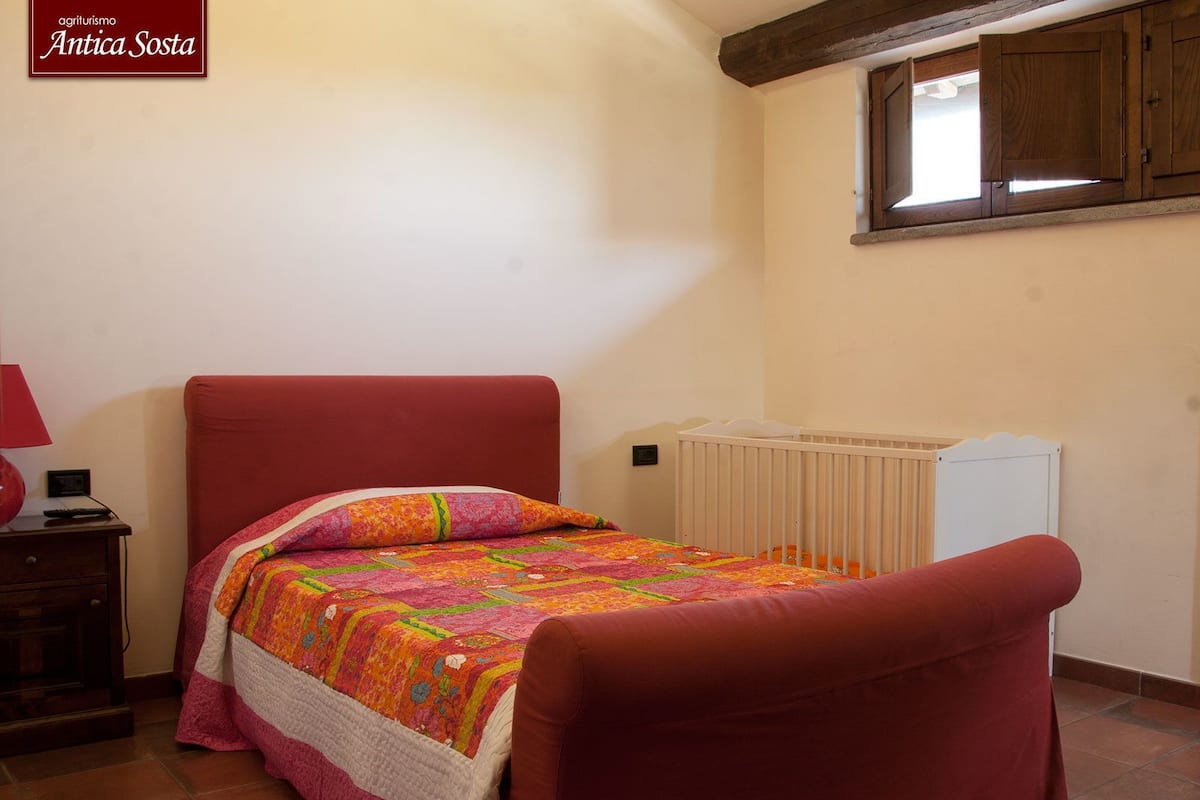 A bedroom with a bed, a nightstand, a lamp, a crib, and a window.