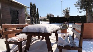 Un banco y una mesa de madera cubiertos de nieve, con una planta en maceta y un edificio al fondo.