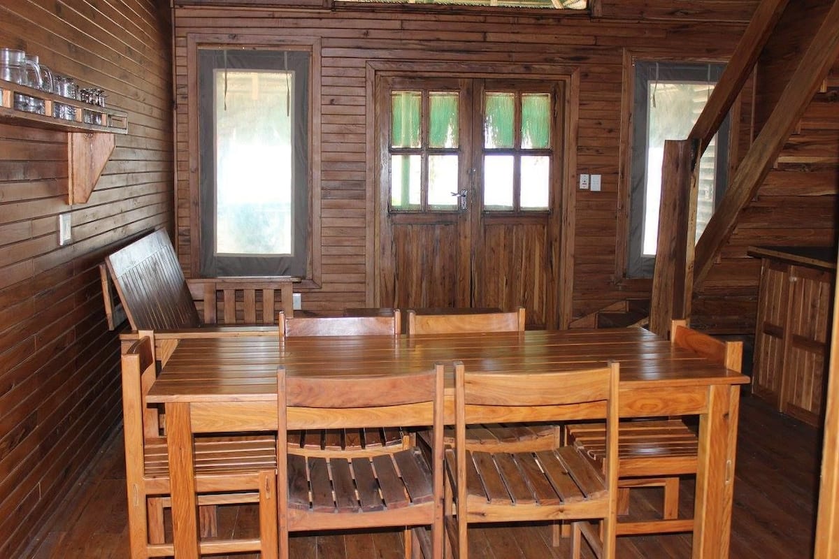 Interior de una cabaña de madera con una mesa y sillas, una ventana con vistas y una puerta con ventanal.