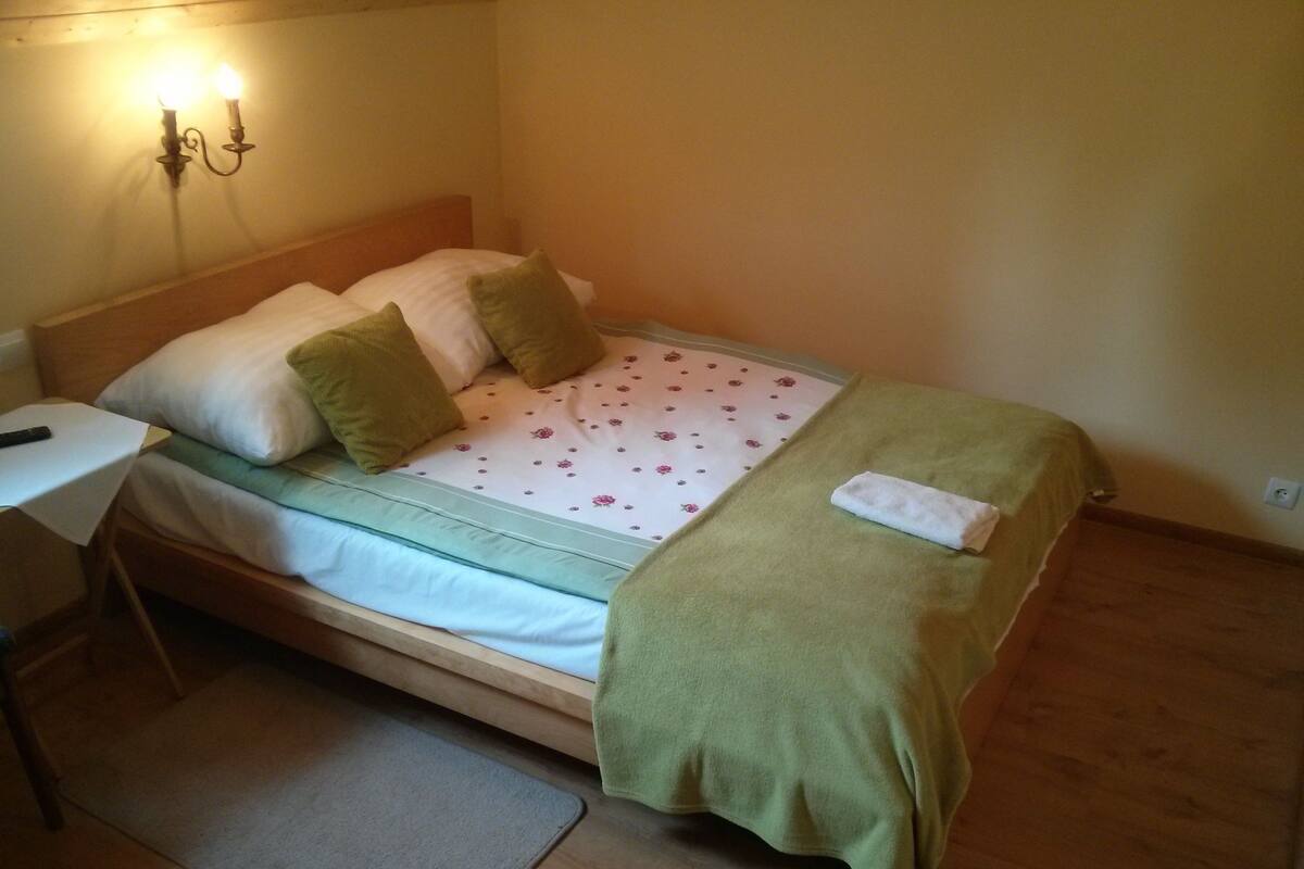 A single bed with a floral bedspread, two pillows, a green blanket, and a bedside table with a lamp.