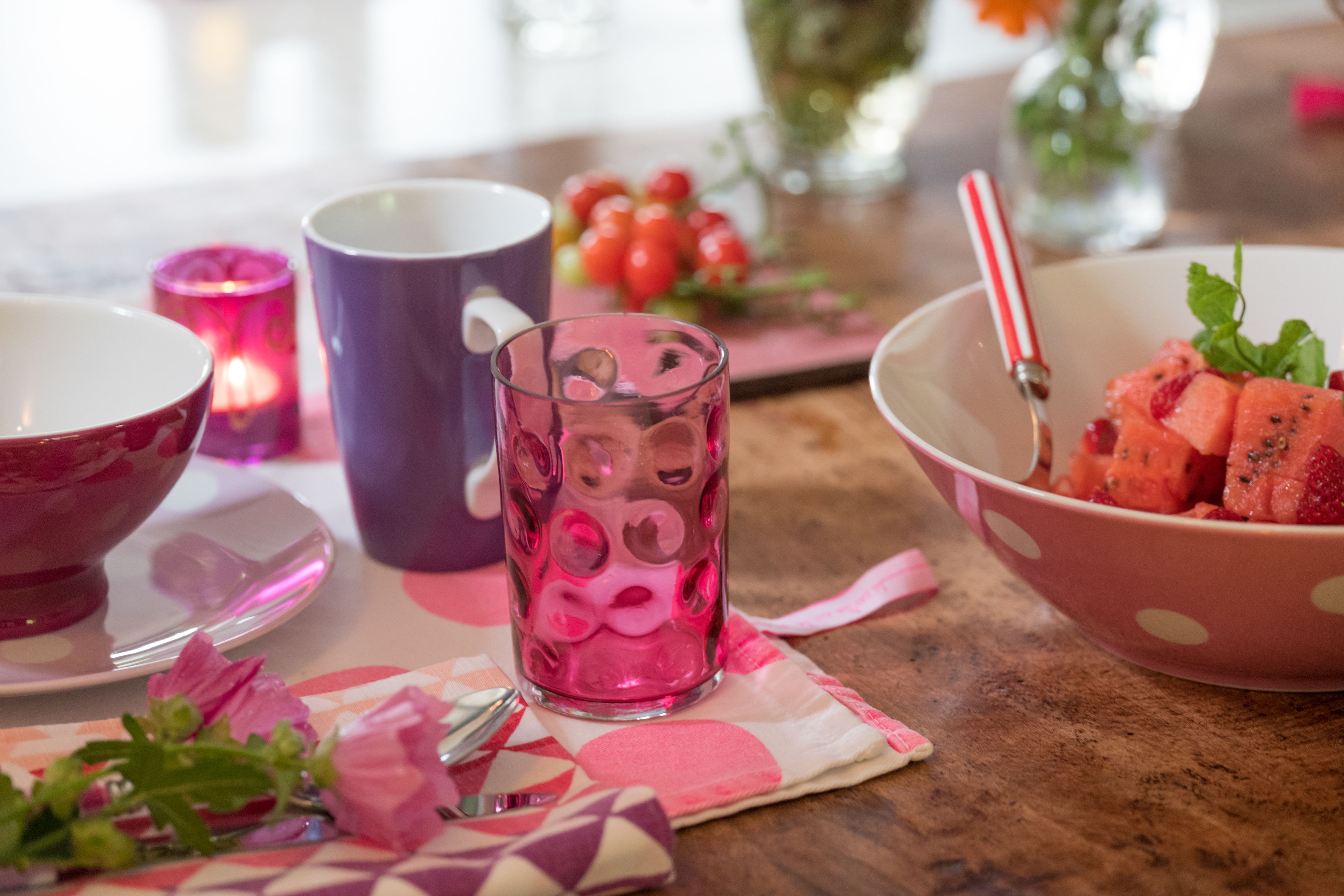 A table with a pink bowl of watermelon, a purple mug, a pink glass, and a candle.