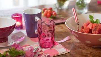 A table with a pink bowl of watermelon, a purple mug, a pink glass, and a candle.