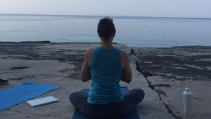 A person sitting on a yoga mat facing the sea, with a notebook and a water bottle nearby. - Lådfabriken (Hälleviksstrand)