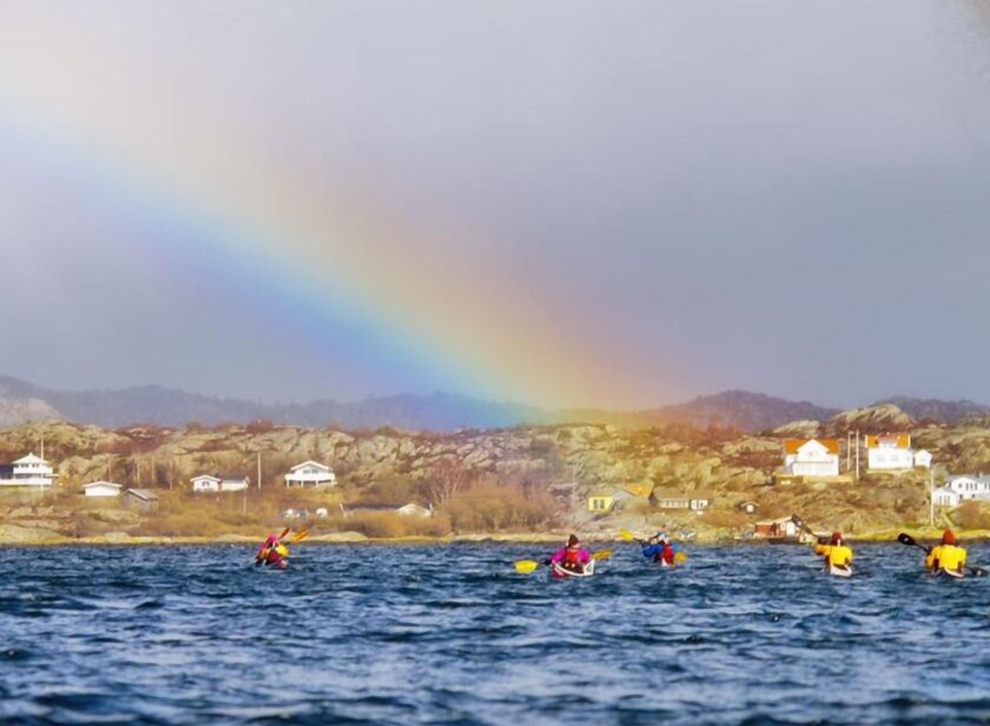 A group of kayakers on a body of water with a rainbow in the background and houses on the shore.