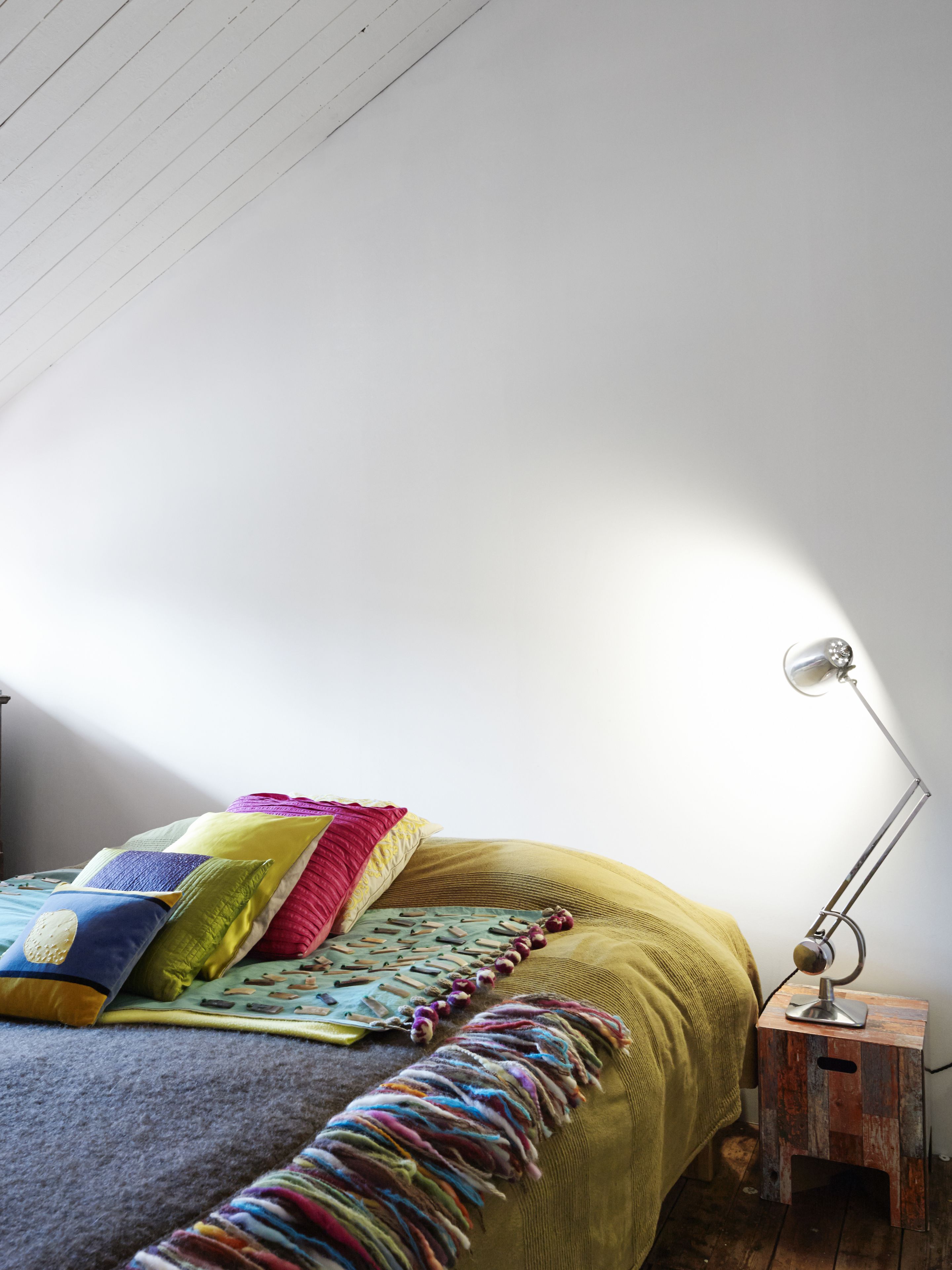 A bedroom with a bed, colorful pillows, a blanket, a lamp, and a small wooden stool.