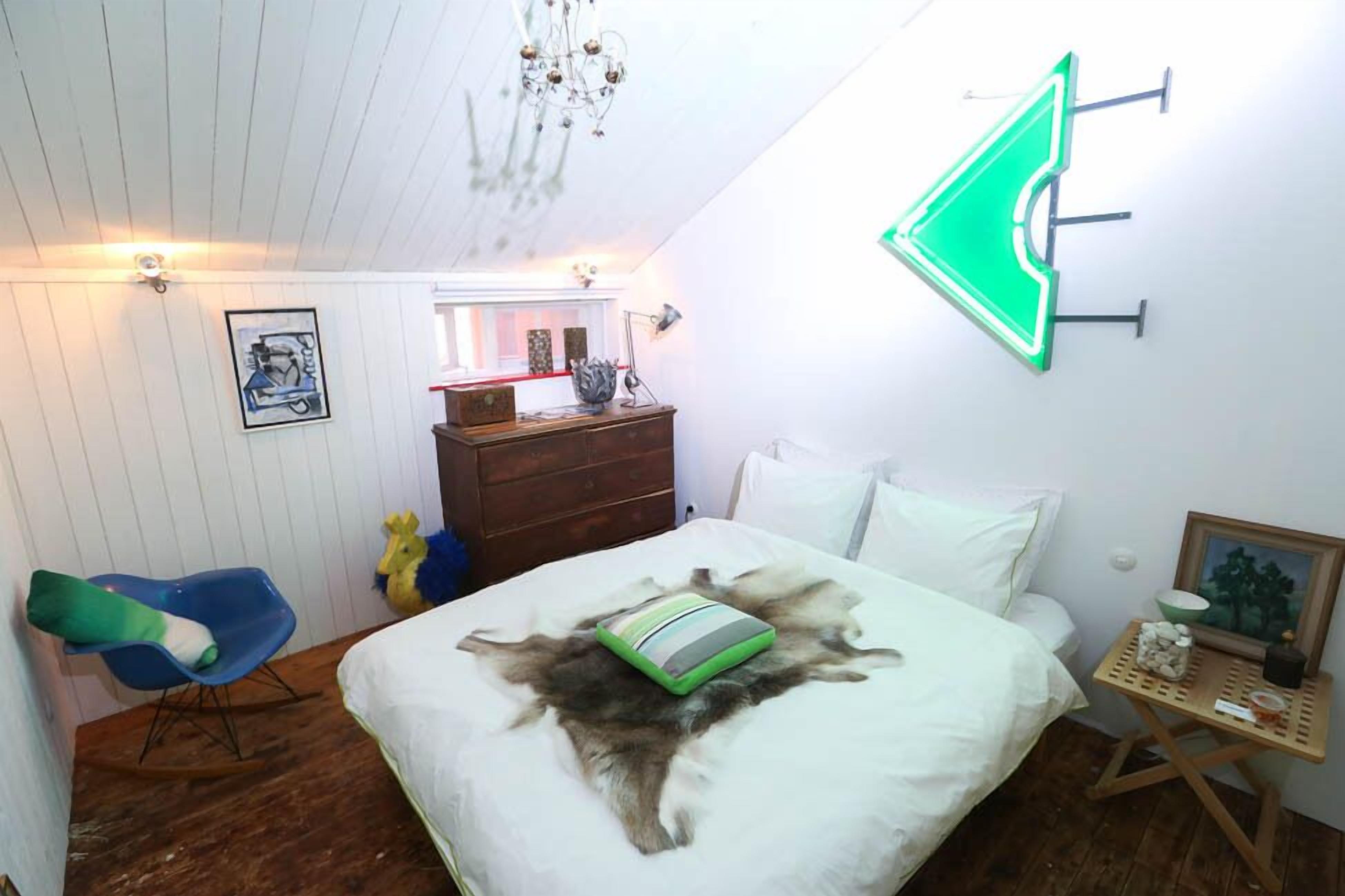 A room with a bed, a dresser, a chair, a neon sign, and a chandelier.