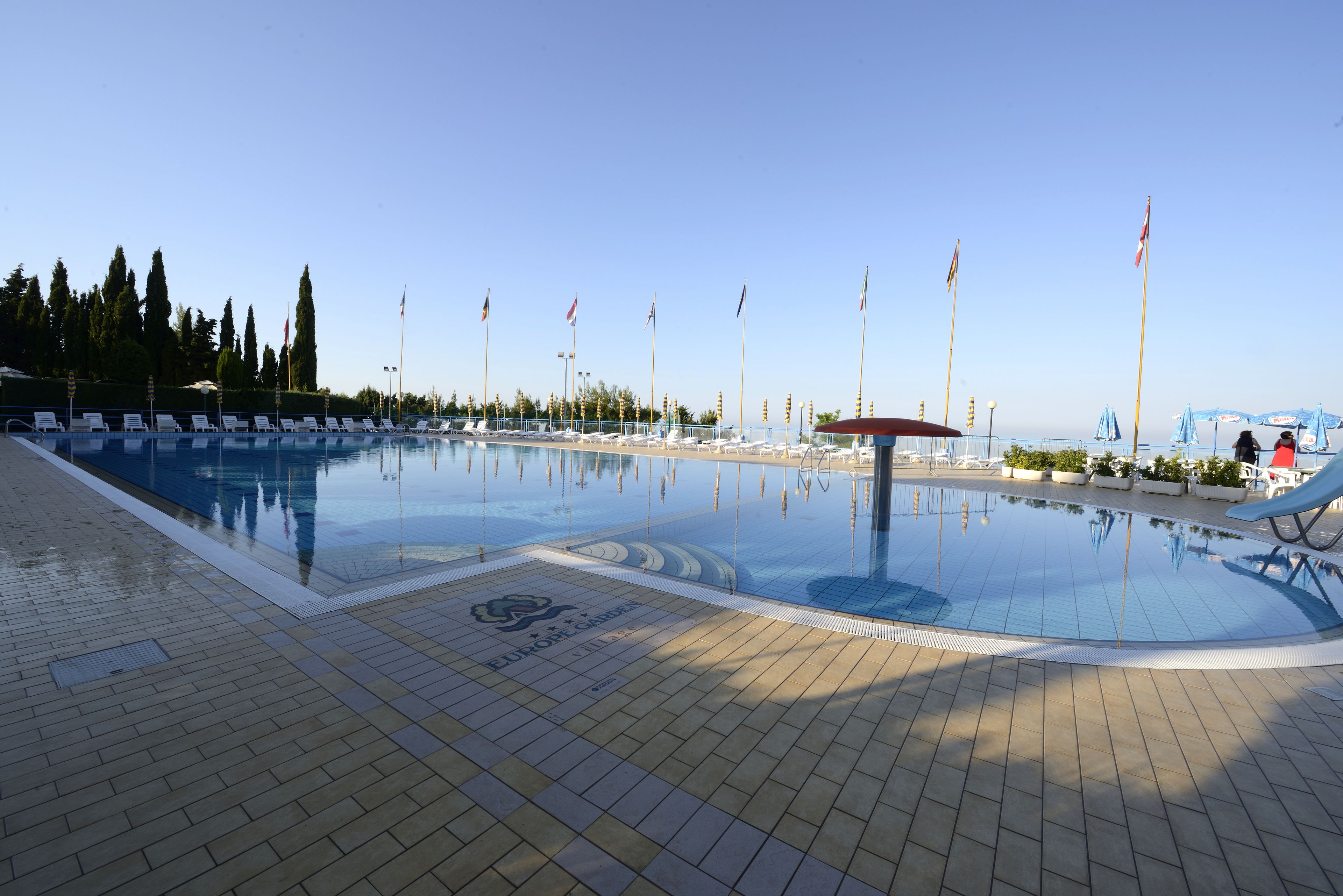 A swimming pool with lounge chairs and umbrellas, surrounded by flag poles and trees.