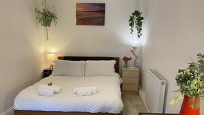 A neatly made bed with white linens and towels, a bedside table with a lamp, a small nightstand with a vase of flowers, and a hanging plant. - Lovely 1 bedroom, with parking & patio, 5 min walk from Lexicon  & Train Station (Bracknell)