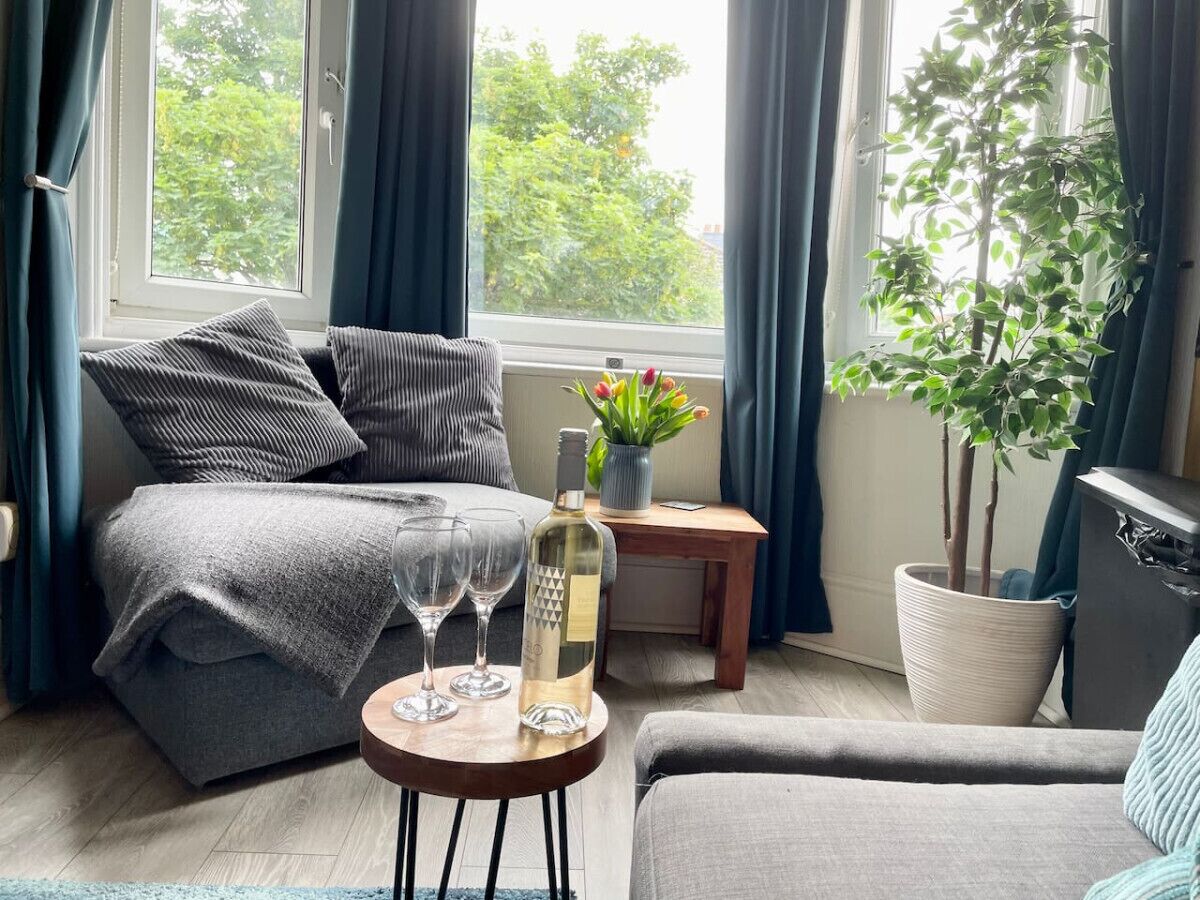A cozy living room with a sofa, a coffee table with glasses and a bottle, a small side table, and a potted plant.