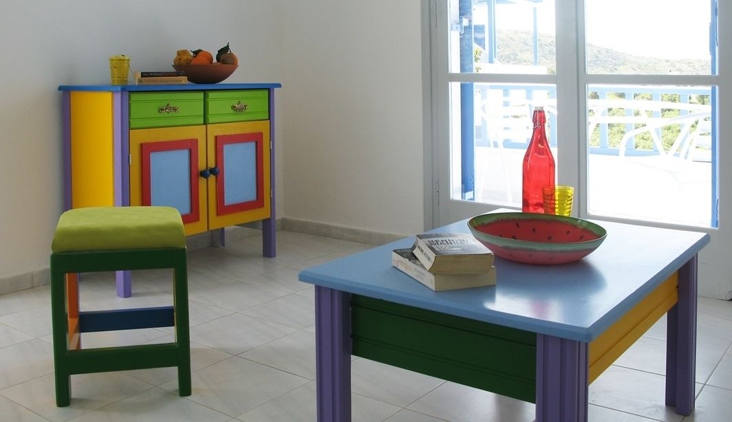 Een kamer met een kleurrijke kast, een groene kruk, een tafel met een kom en een boek, en een raam met uitzicht op een witte schutting en begroeiing.