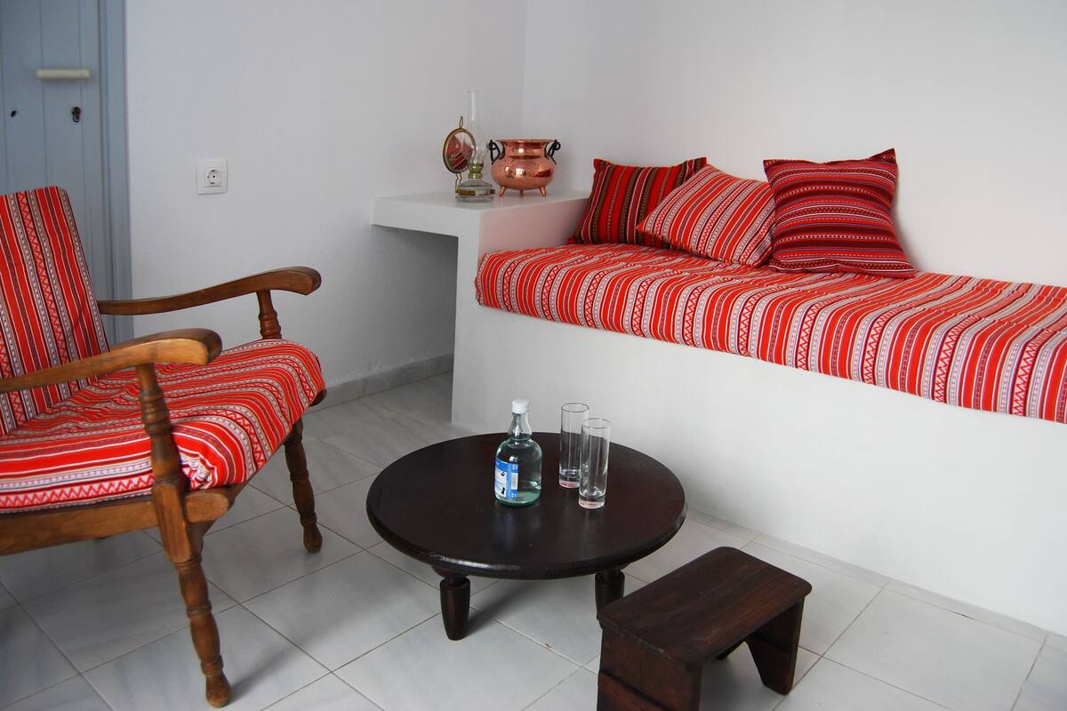 Una habitación acogedora con una silla y sofá de madera, una mesita con una botella y vasos, y una olla de cobre decorativa en una estantería.