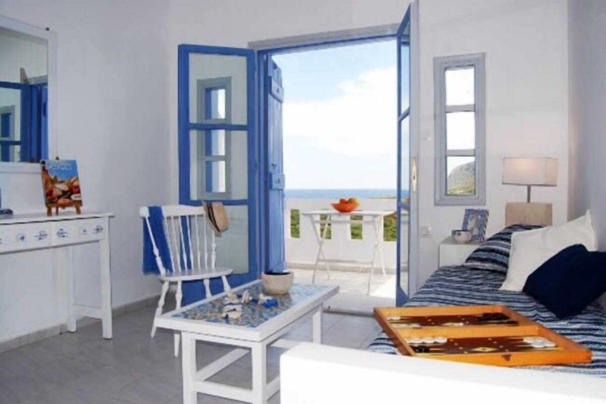 Una habitación con cama, una mesa, una silla y vista al mar por una puerta abierta.