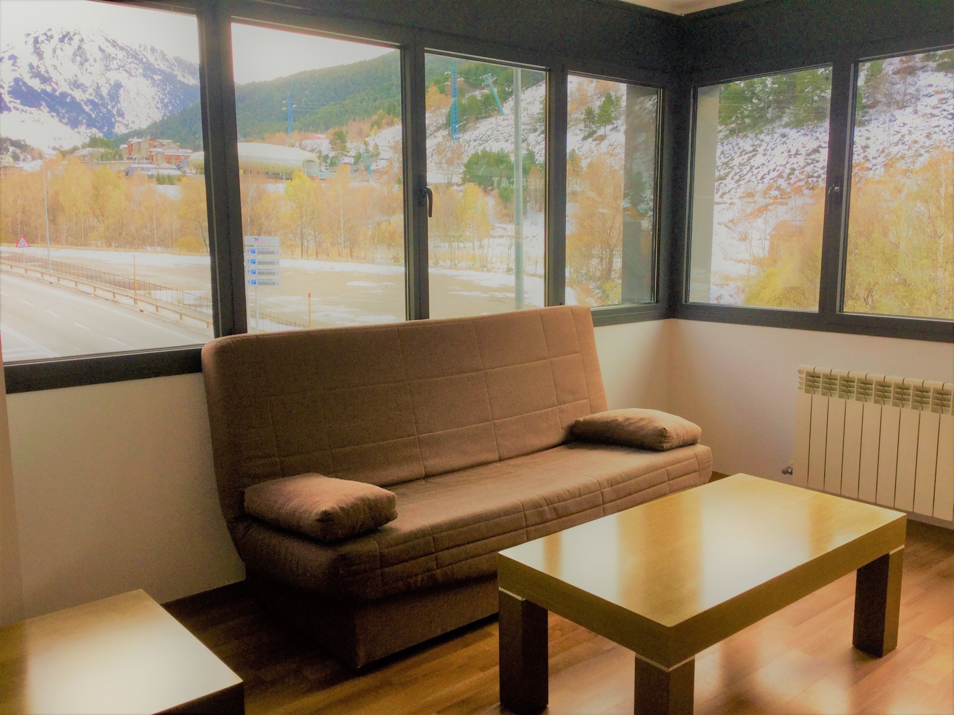 A room with a brown sofa, a wooden table, and large windows offering a view of a snowy landscape and mountains.