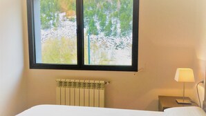 Una habitación con cama, ventana con vista a árboles, radiador y mesita de noche con lámpara.