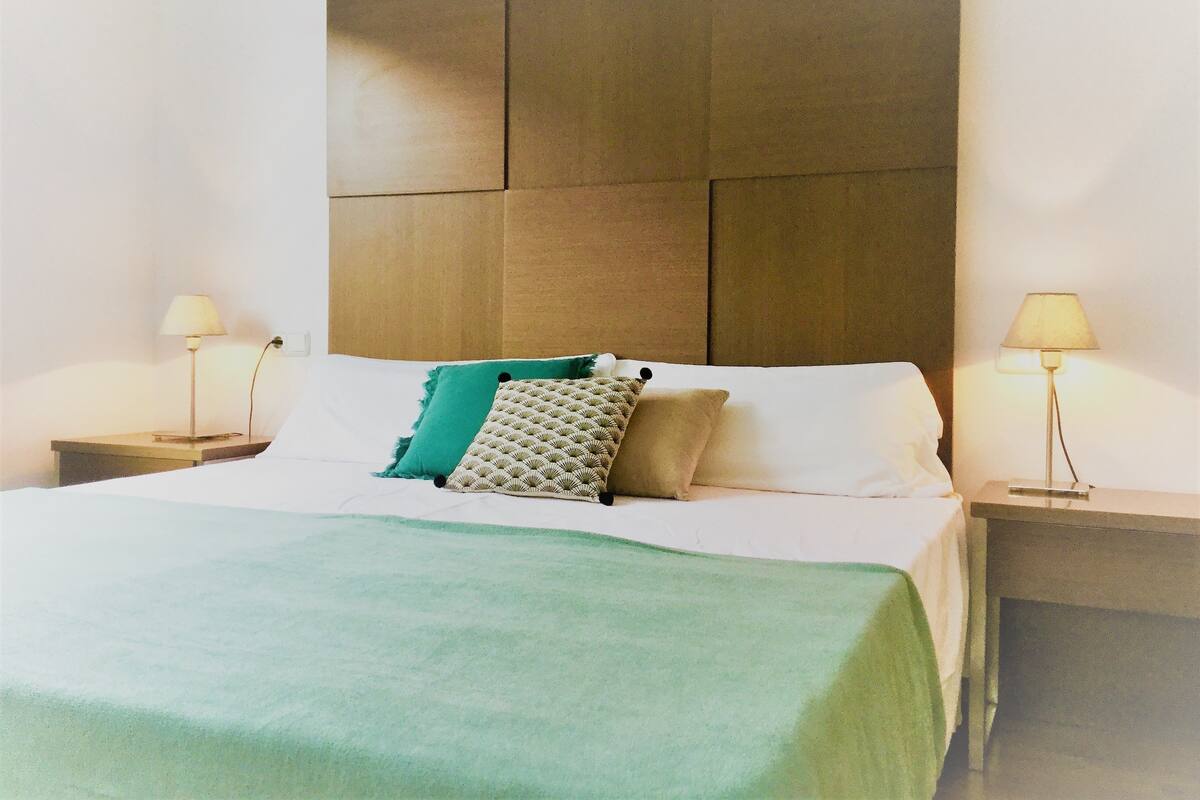 A neatly made bed with a green throw blanket, two pillows, and a wooden headboard.