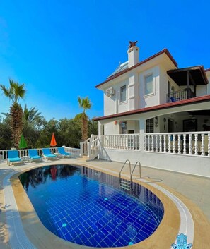 A swimming pool with a white balustrade, a two-story building with a red roof, and palm trees in the background.