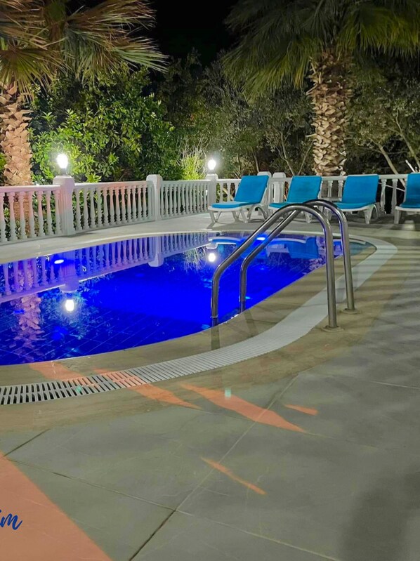 Une piscine extérieure entourée de palmiers et de verdure, avec une clôture blanche et des chaises bleues.