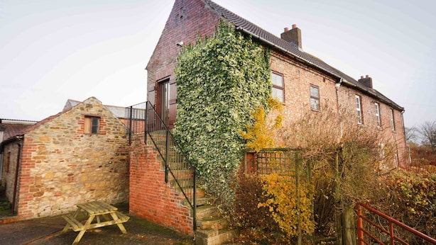 A traditional brick building with a steeply pitched roof, a stone structure to the left, and a wooden bench in the foreground. - The Granary at Todds House Farm  (Stockton on Tees)