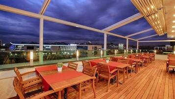 Ein Dachrestaurant mit Holztischen und -stühlen, einer Glasufernung und Blick auf die Skyline der Stadt im Abendrot.