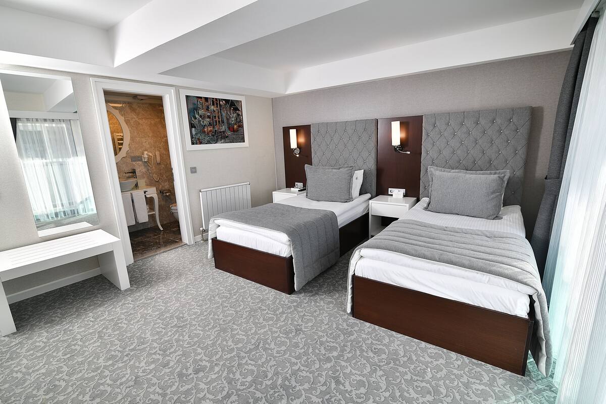 A modern hotel room with two beds, a bench, a bathroom, and a view of the city.