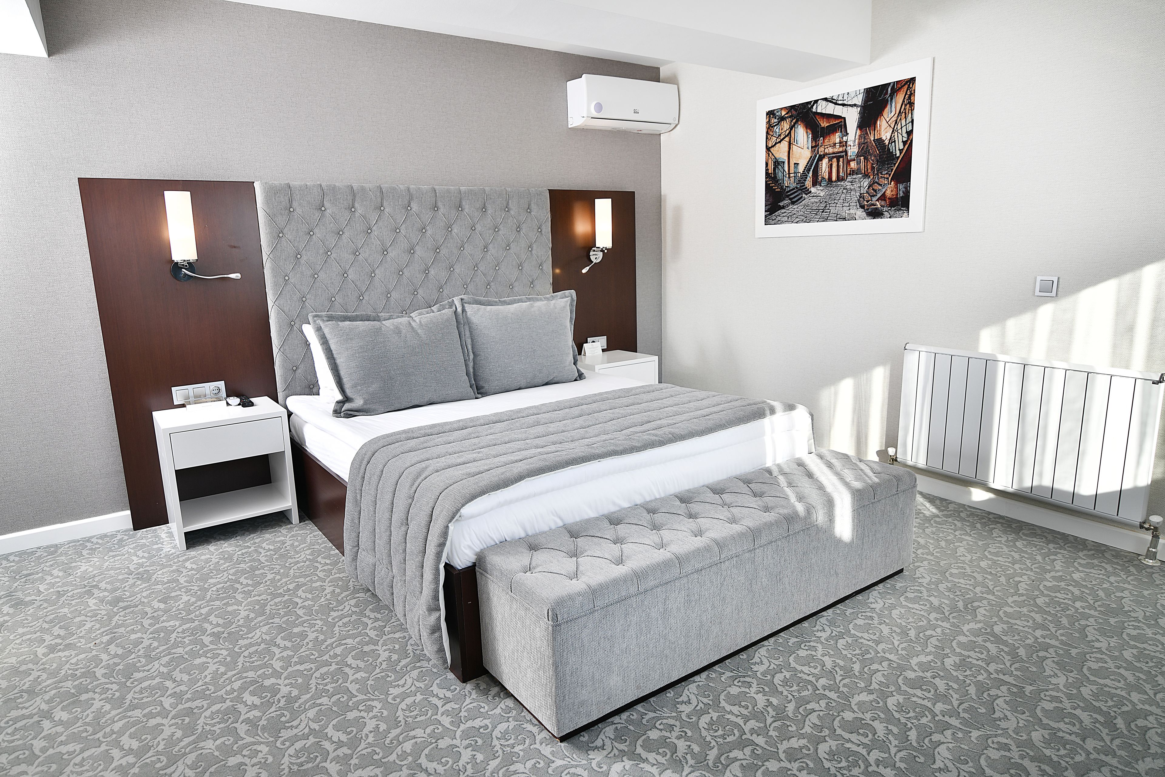 Un dormitorio moderno con una cabecera capitonada, una mesita de noche blanca, un banco gris y una alfombra estampada.