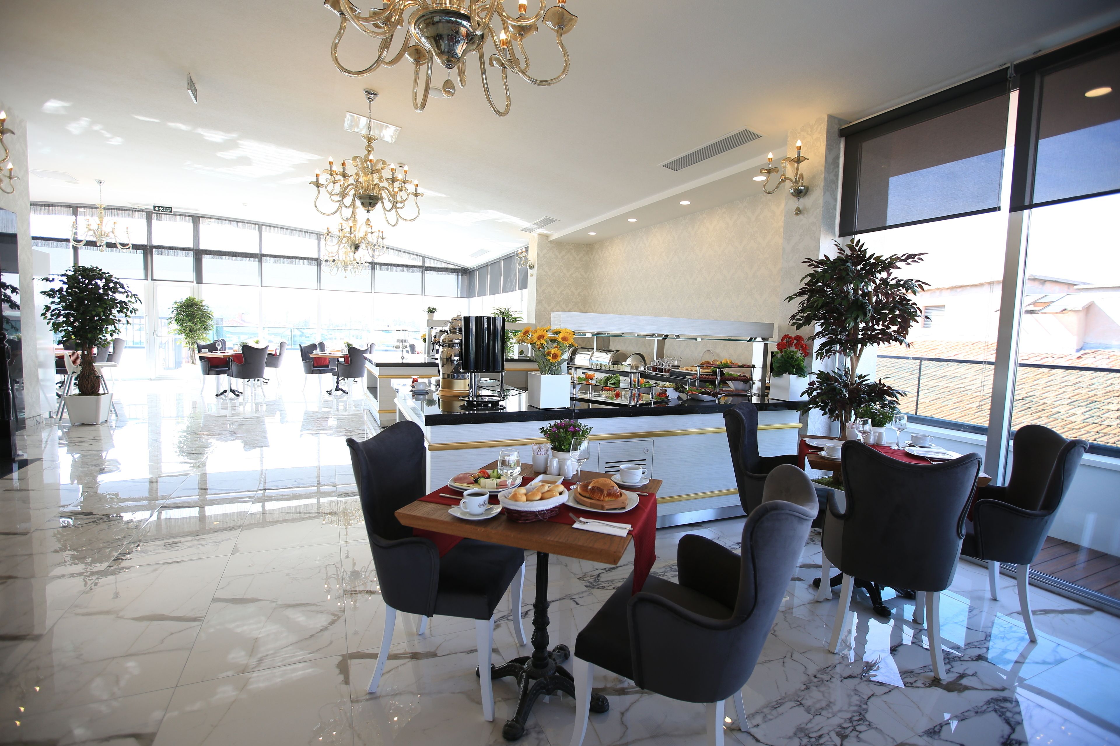 Ein moderner Hotel-Speiseraum mit Marmorboden, schwarzen und grauen Stühlen, einem Holztisch mit Frühstücksangeboten und großen Fenstern mit Blick.