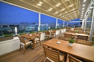 Un patio al aire libre con mesas y sillas de madera, plantas en macetas y vistas a la ciudad al atardecer.