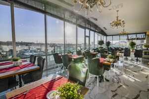Un comedor moderno con grandes ventanales, varias mesas y sillas, y vistas a la ciudad.