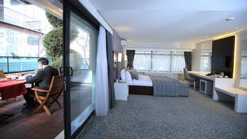 A modern hotel room with a balcony, a bed with grey bedding, a desk with a chair, and a television.