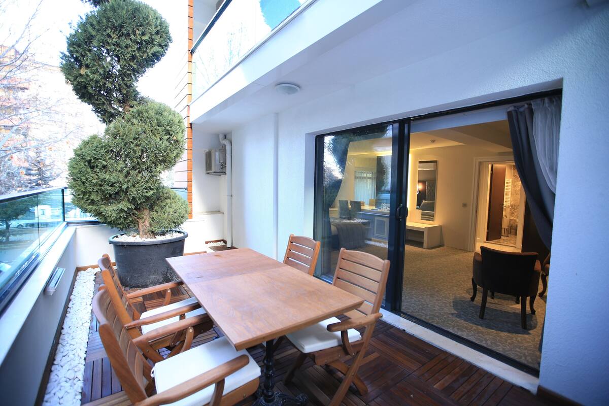 A balcony with a wooden table and chairs, a potted tree, and a view of the interior through a glass door.