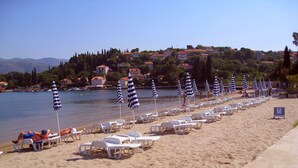 Una spiaggia con file di poltroncine bianche e ombrelloni a righe, mare calmo e edifici sullo sfondo.