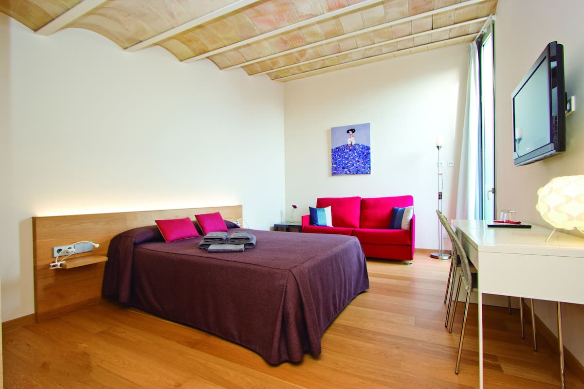 A bedroom with a bed, a desk, a chair, a red sofa, and a painting on the wall.