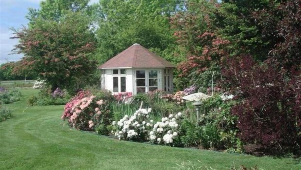 Un petit kiosque blanc, surmonté d’un toit rouge, est entouré d’une végétation luxuriante et de plantes en fleurs.