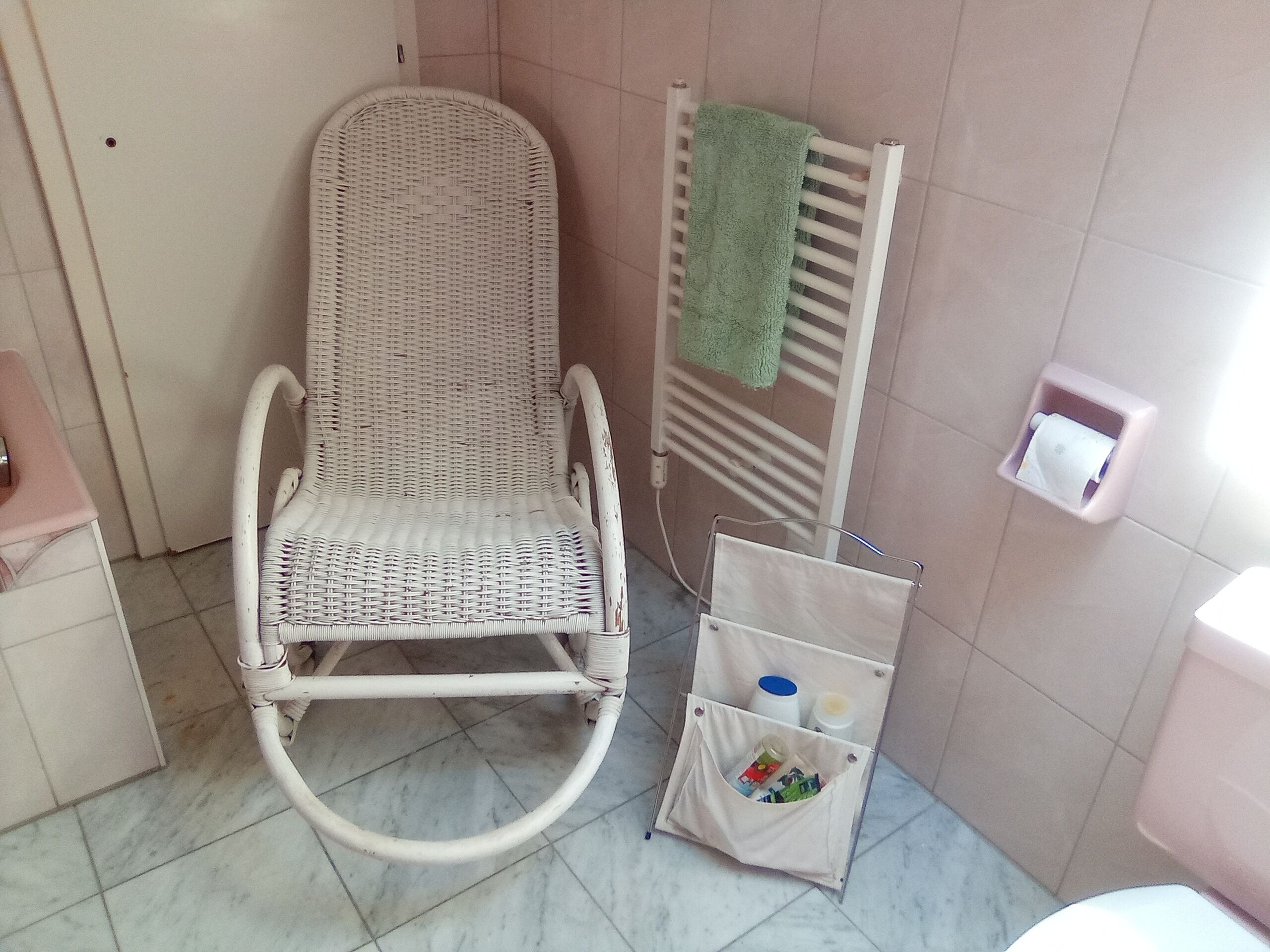 Un baño con una silla de mimbre blanca, un perchero con una toalla verde, un secador de manos y un pequeño cofre para guardar objetos.