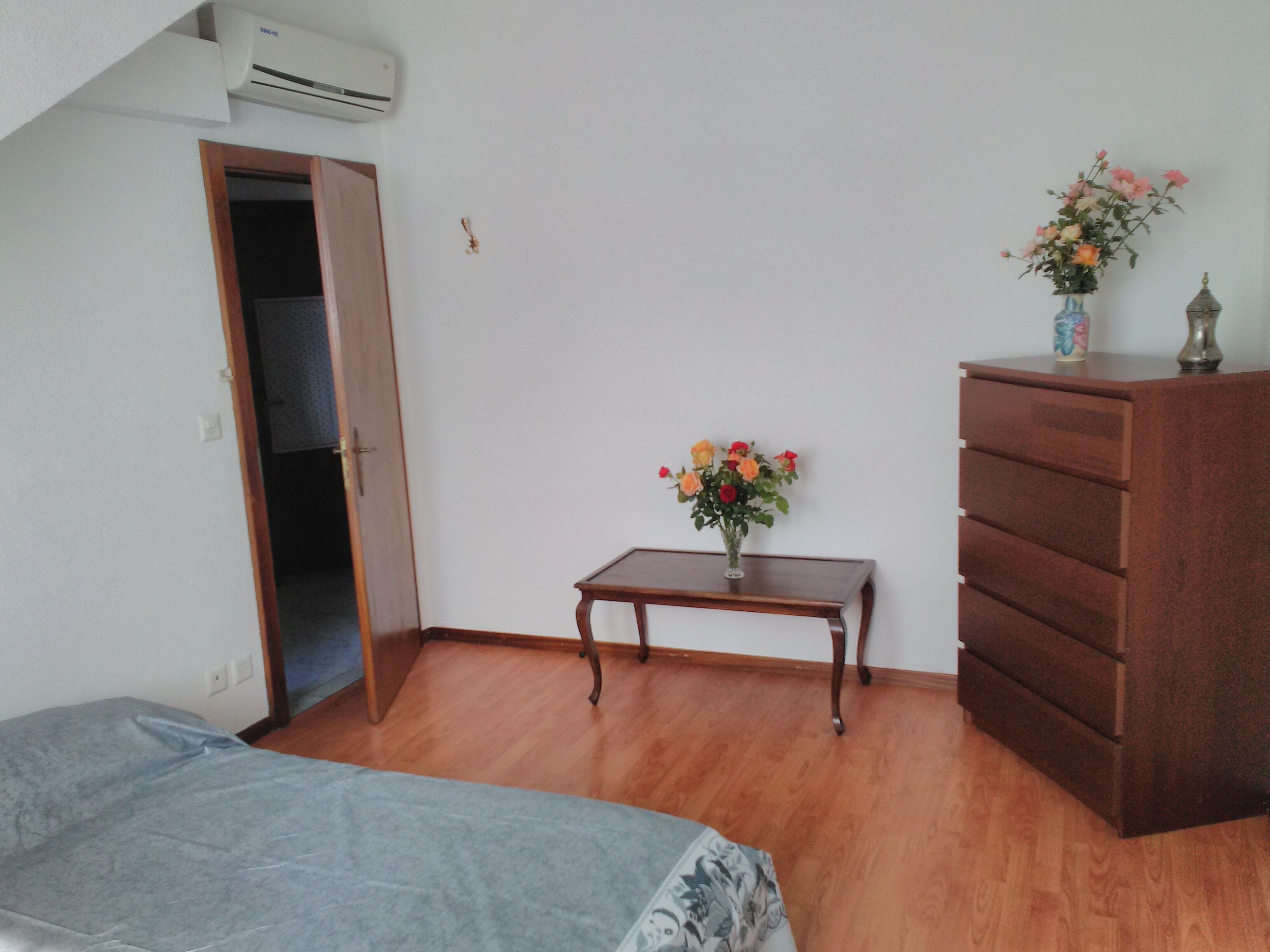 Ein Schlafzimmer mit Bett, Kommode, einem kleinen Tisch mit Blumen und einer Wand-Klimaanlage.