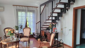 Un vaste salon agrémenté d’un escalier en bois, un coin repas avec une table ronde et des chaises, et une vue sur l’extérieur grâce à de grandes fenêtres.