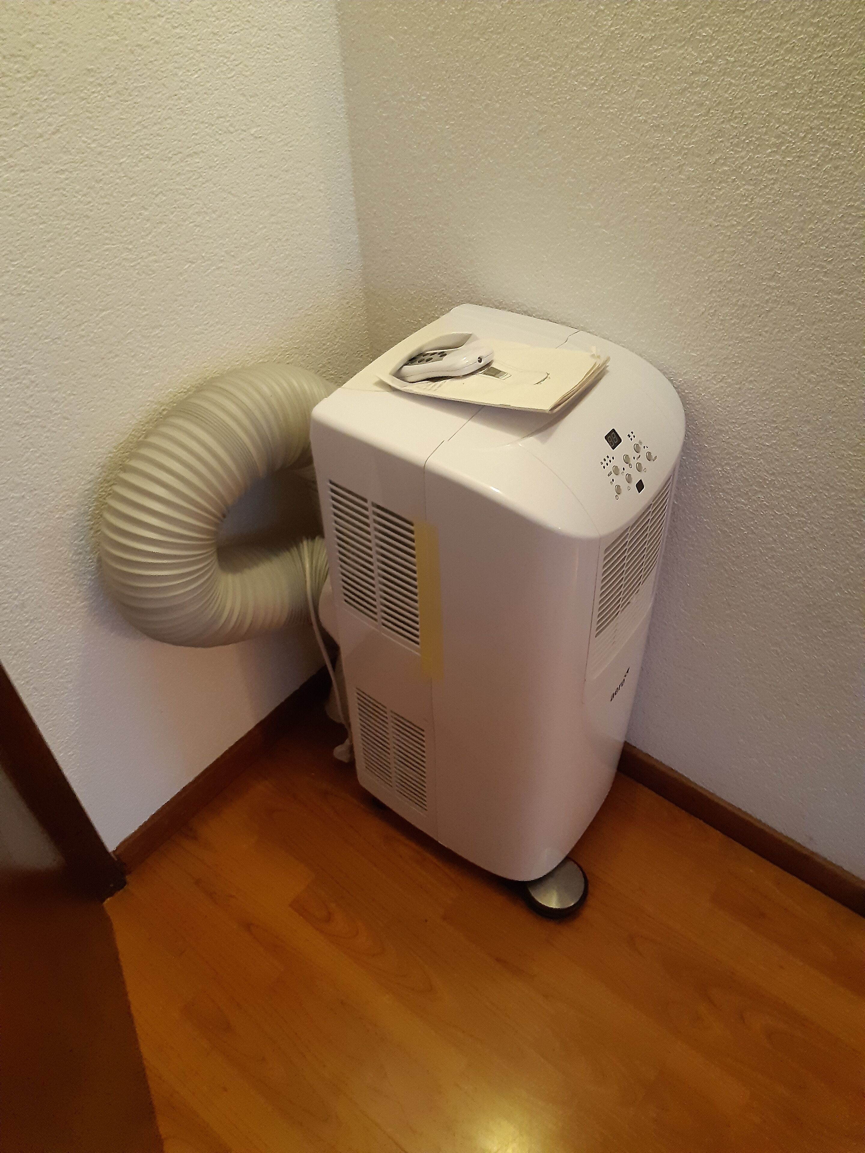 Ein weißer, tragbarer Heizlüfter mit Bedienfeld und flexiblem Abluftschlauch, platziert in einer Ecke eines Raumes mit Holzboden und weißen Wänden.