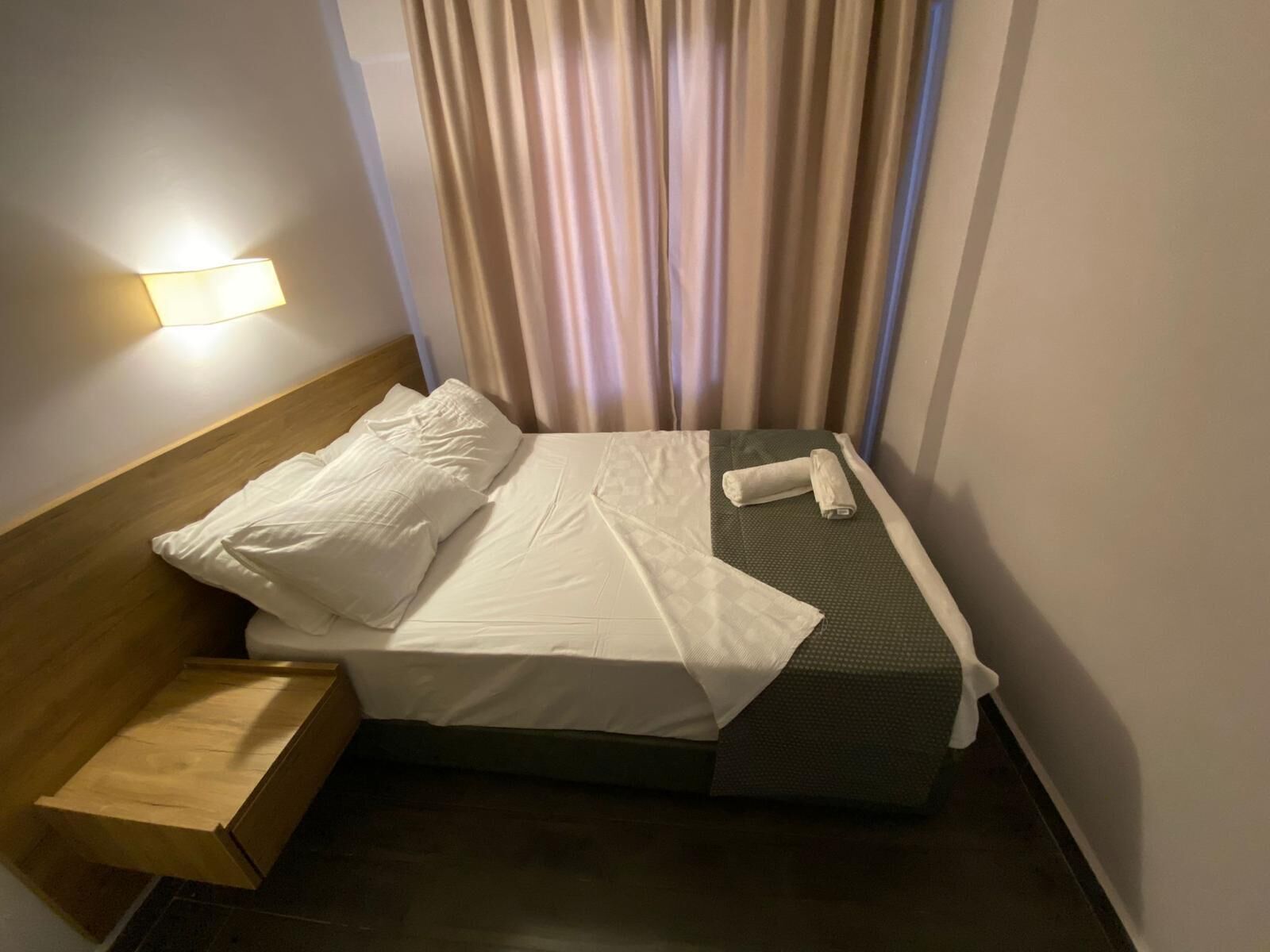 A neatly made bed with white linens, a wooden headboard, a small wooden nightstand, and a wall-mounted light.