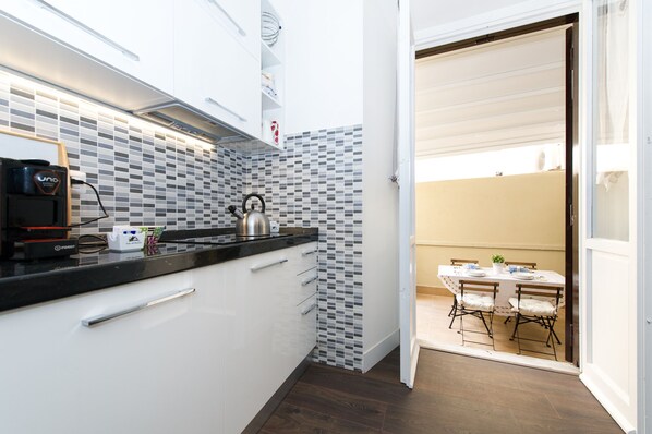 A compact kitchen with white cabinets, a black countertop, and a tiled backsplash. There's a kettle on the counter and a coffee machine. A dining area with a table and chairs is visible through an open door.