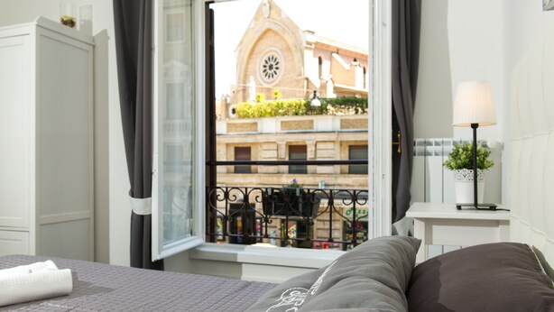 Habitación de hotel con cama, ventana que ofrece vistas a un edificio con un reloj, una mesita con lámpara y una planta en el alféizar.