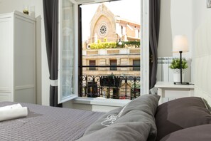 A hotel room with a bed, a window with a view of a building with a clock, a small table with a lamp, and a plant on the windowsill.