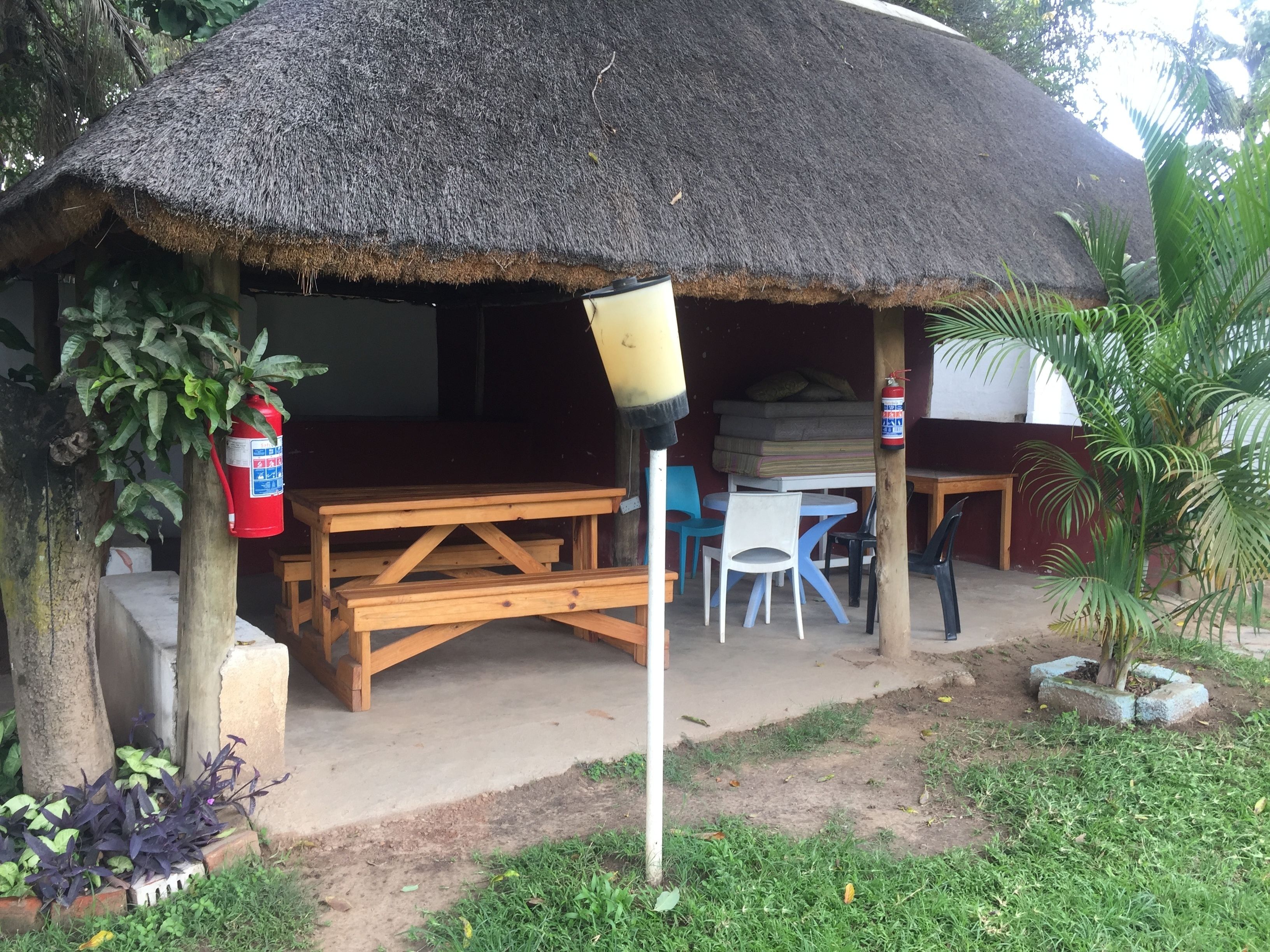 A thatched-roof hut with a wooden table and chairs, a fire extinguisher, and a small garden area.
