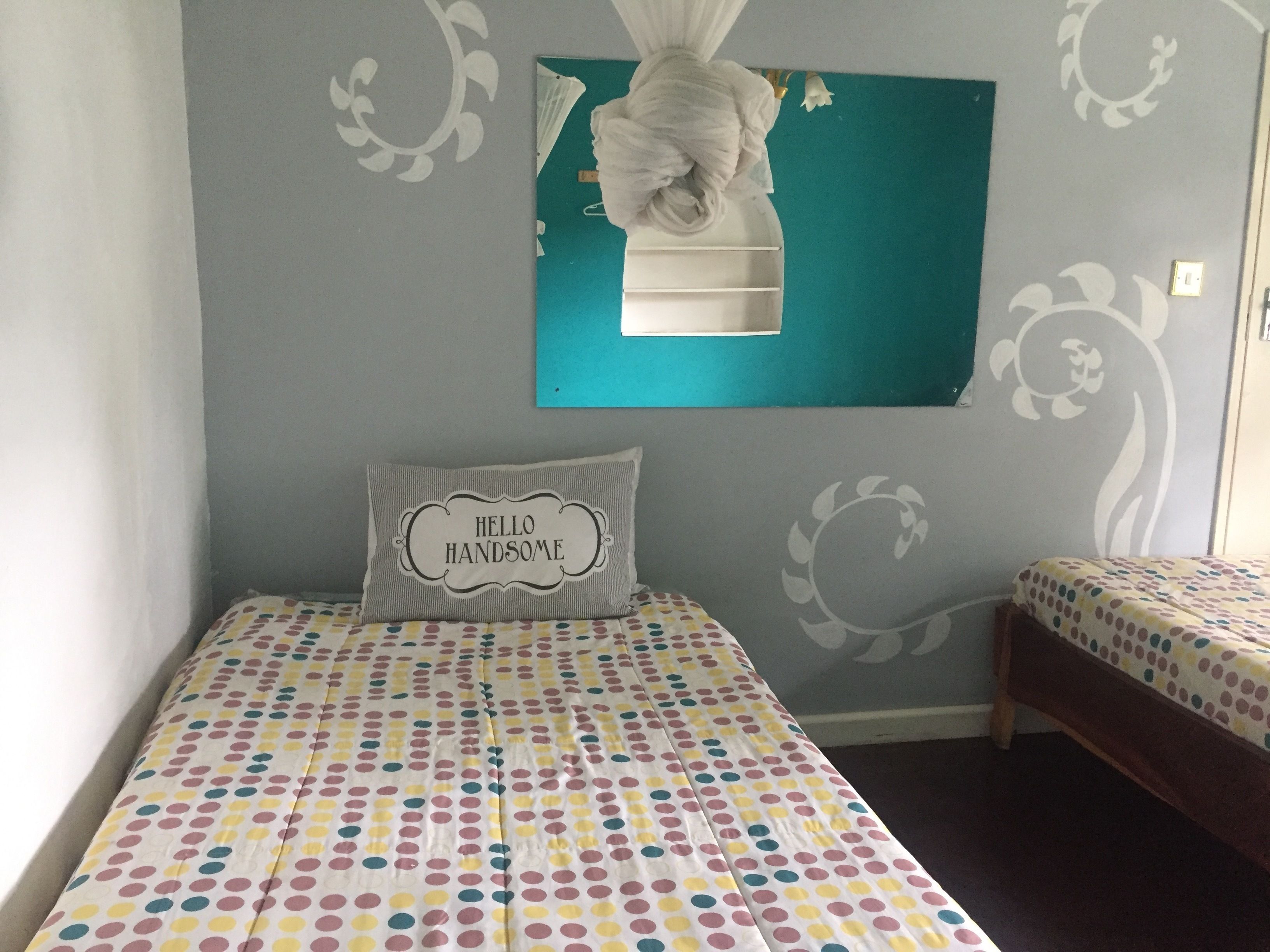 A bedroom with a bed featuring a polka dot bedspread, a pillow with "HELLO HANDSOME" text, and a wall with a floral design.