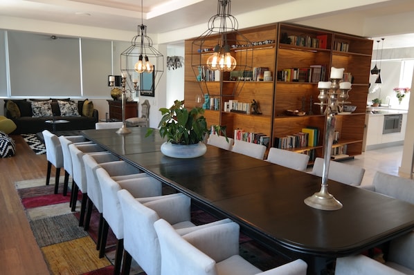 A modern dining area with a long table, white chairs, a central plant, and a large bookshelf filled with books.