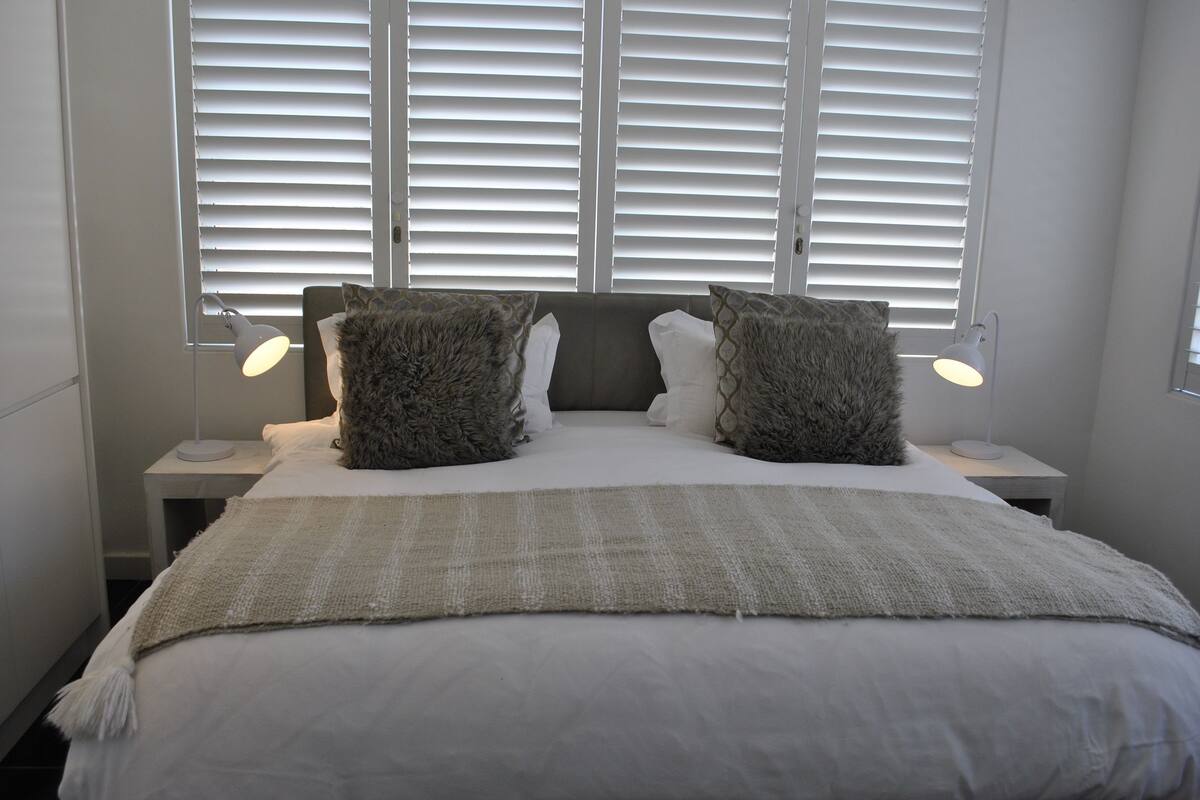 A neatly made bed with a grey and white color scheme, two bedside lamps, and a window with blinds.