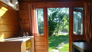 A wooden cabin with a kitchen area, a dining table, and a view of a grassy yard through the open door. - Camping Baalse Hei (Turnhout)