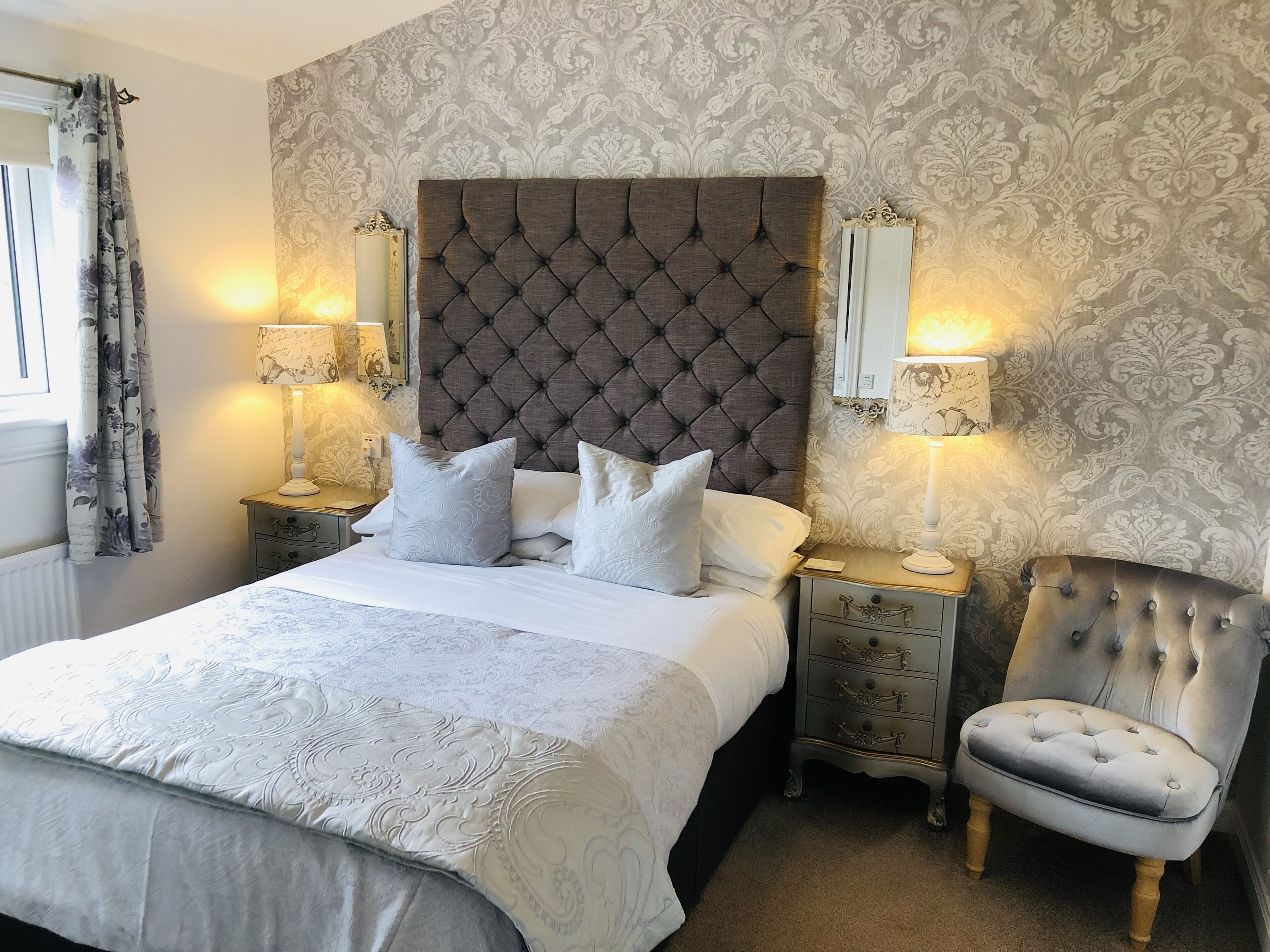 A neatly made bed with a tufted headboard, flanked by two bedside tables with lamps, a chair, and patterned wallpaper.