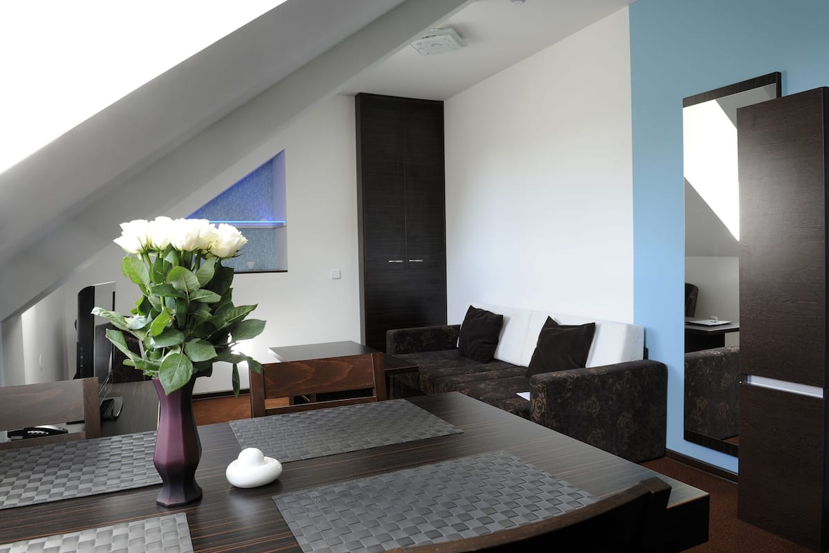 Un salon moderne avec un ensemble de canapés marron foncé, une table à manger en bois agrémentée de sets de table, un vase avec des fleurs blanches et une grande fenêtre laissant entrer la lumière naturelle.
